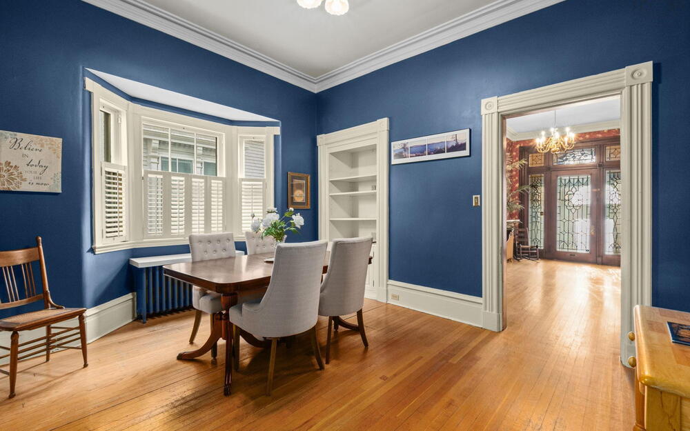 Hardwood flooring, bay window with plantation shutters, crown molding, built-in shelving, and fluted door casings